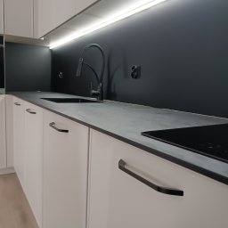 Maciej Kukuła - Nowoczesna, minimalistyczna kuchnia w Gorlicach: białe szafki z czarnymi uchwytami, szary blat, czarny panel nad blatem i oświetlenie LED pod szafkami wiszącymi.