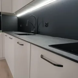 Nowoczesna, minimalistyczna kuchnia w Gorlicach: białe szafki z czarnymi uchwytami, szary blat, czarny panel nad blatem i oświetlenie LED pod szafkami wiszącymi.