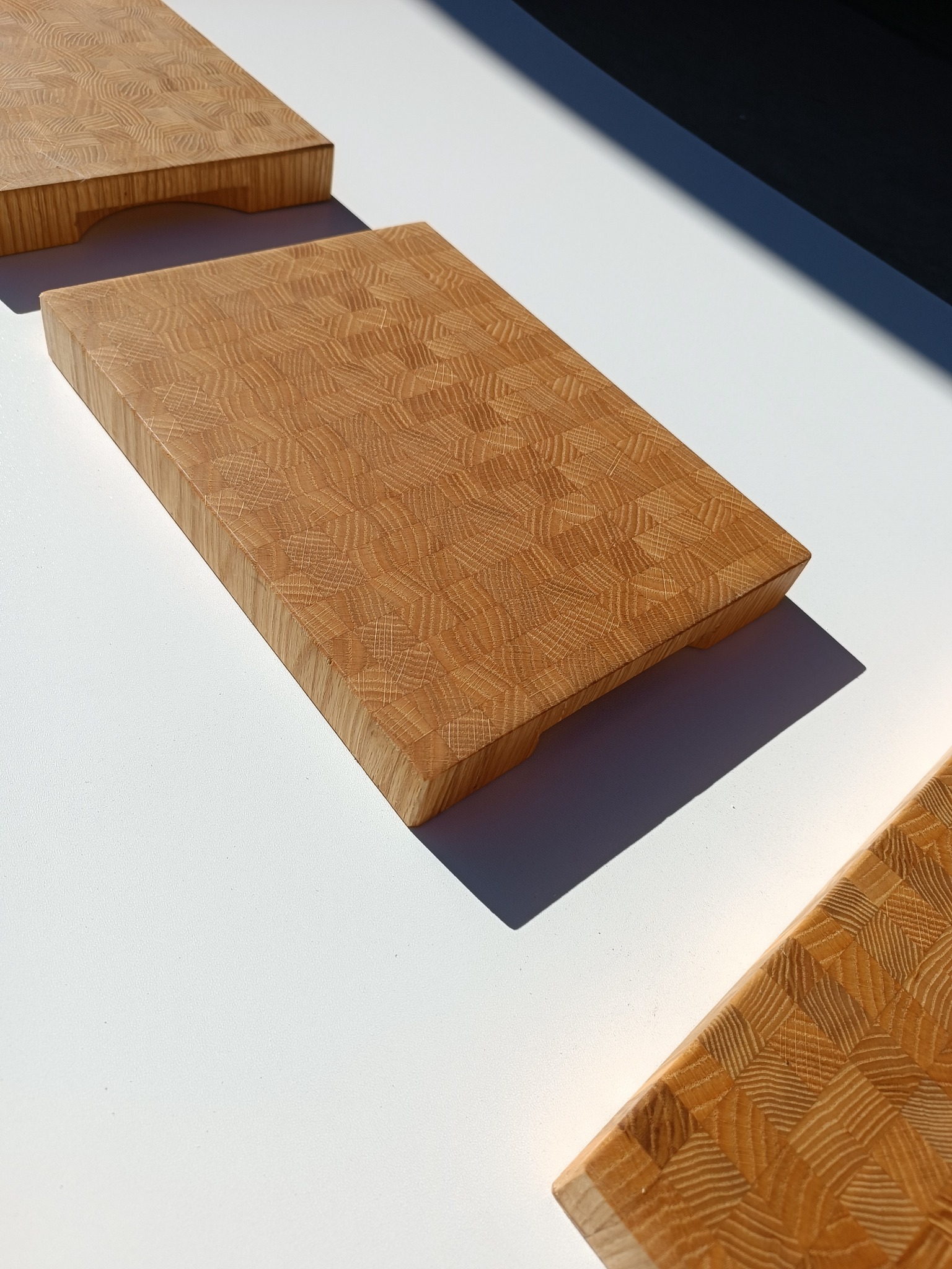 Dębowe deski do krojenia z mozaikowym wzorem, ułożone na białym blacie w jasnym świetle słonecznym, widoczne cienie.