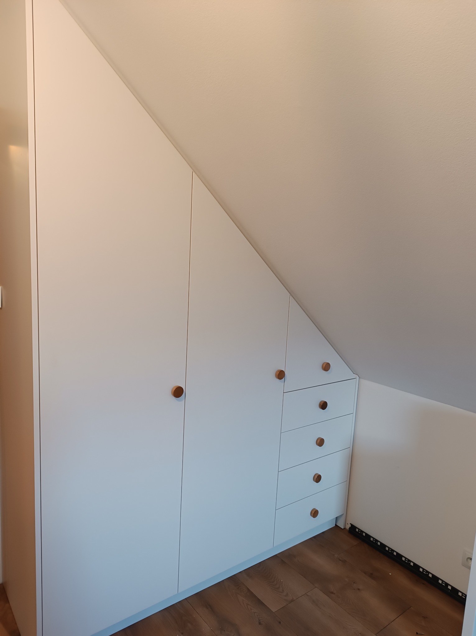 Biała zabudowa meblowa pod skosem na poddaszu, z frontami na dotyk i okrągłymi, drewnianymi uchwytami, dopasowana do kąta nachylenia dachu, z widoczną podłogą z paneli imitujących drewno.