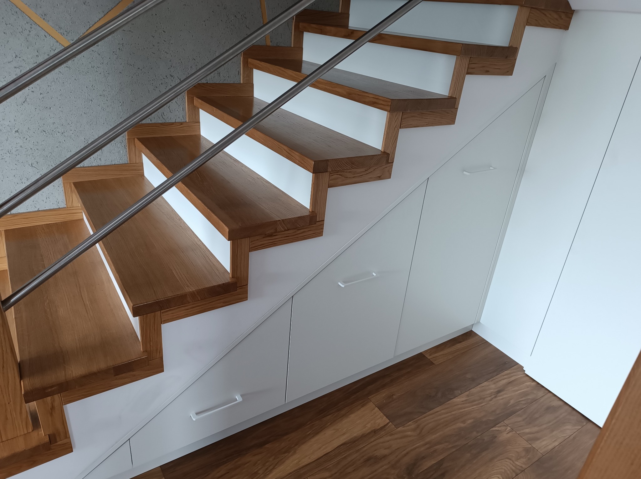 Drewniane schody z białymi podstopnicami i metalową balustradą, zabudowane szafkami z białymi frontami i podłużnymi uchwytami, na podłodze panele drewnopodobne.