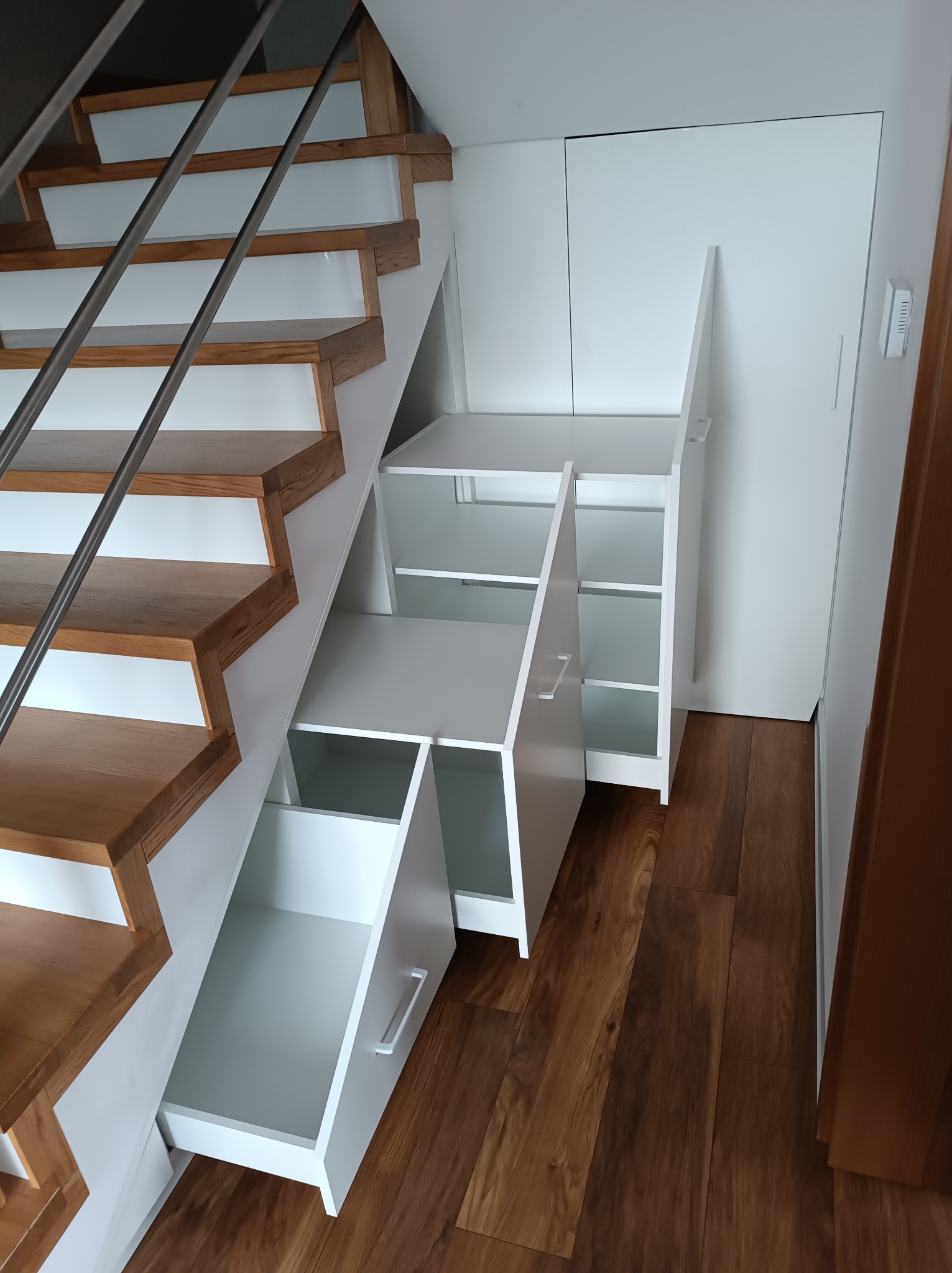 Białe szafki i szuflady w zabudowie pod schodami z drewnianymi stopniami i metalową barierką, widoczne otwarte półki i wysunięta szuflada.