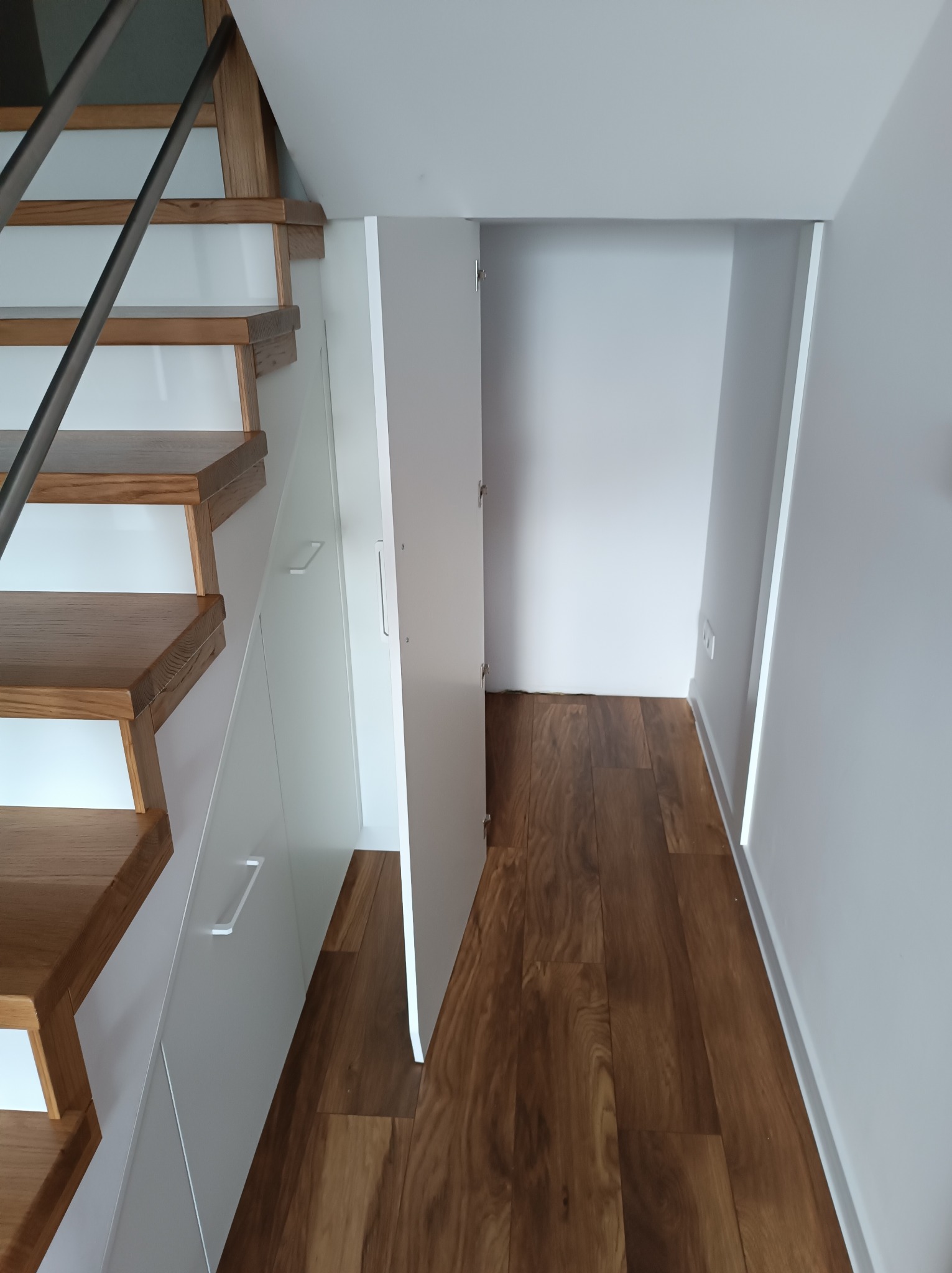 Schody drewniane z zabudową szaf wnękowych pod nimi, białe fronty szafek, otwarte drzwi do przestrzeni schowka, parkiet drewniany.