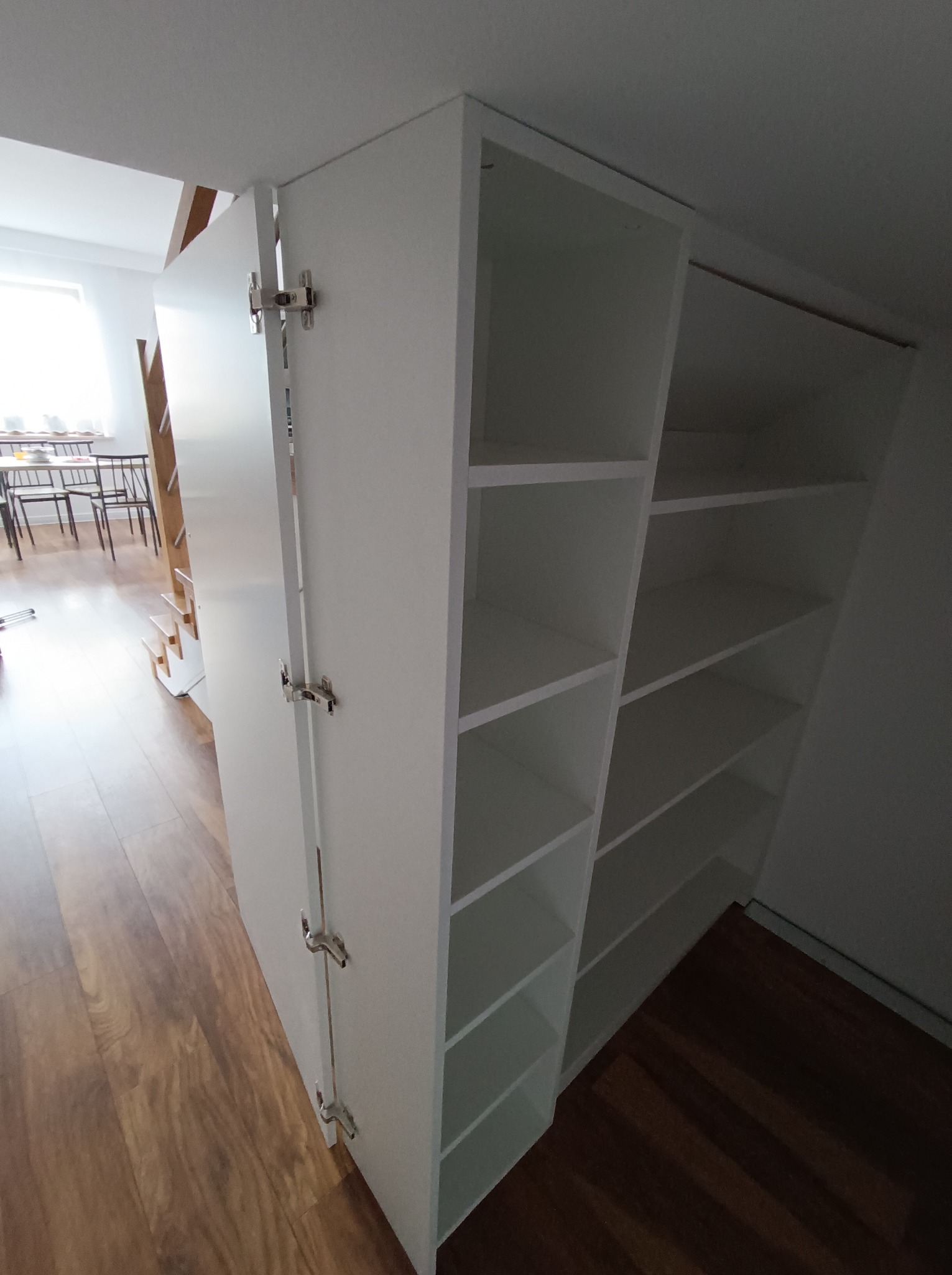Biała szafa wnękowa z otwartymi półkami, zamontowana na poddaszu, widoczne metalowe zawiasy drzwi i drewniana podłoga.