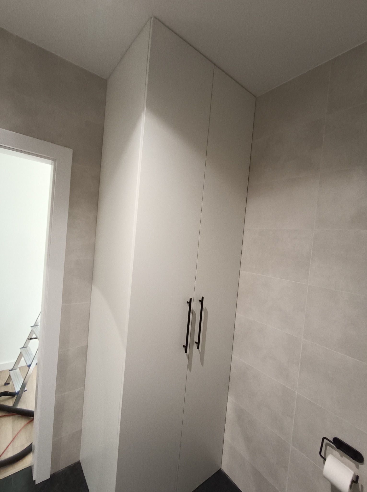 Wysoka, biała szafa z czarnymi uchwytami w łazience wyłożonej szarymi płytkami; w tle widoczny fragment drabiny.