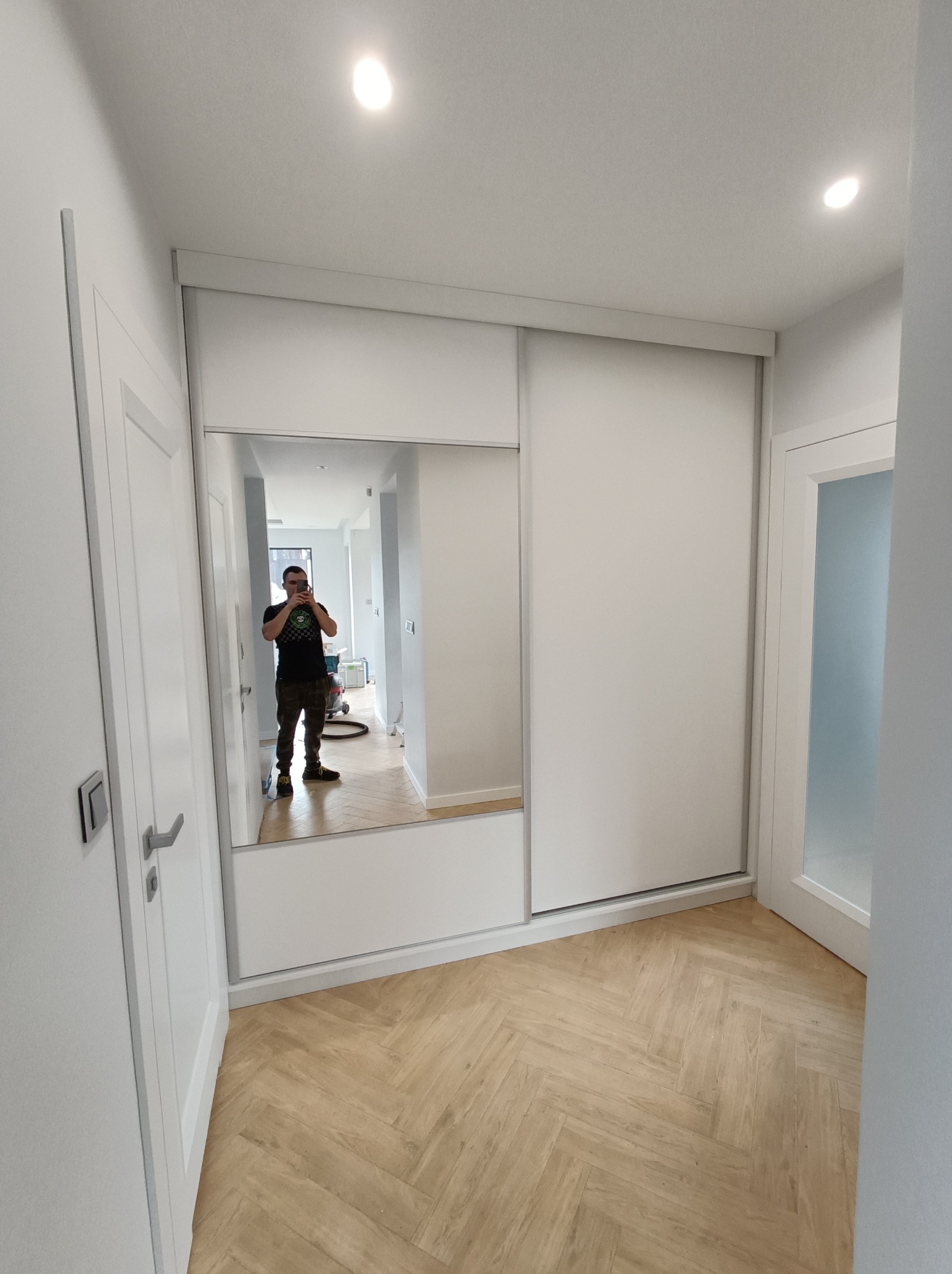Biała szafa przesuwna z lustrem w przedpokoju, widoczny parkiet w jodełkę i odbicie fotografa.