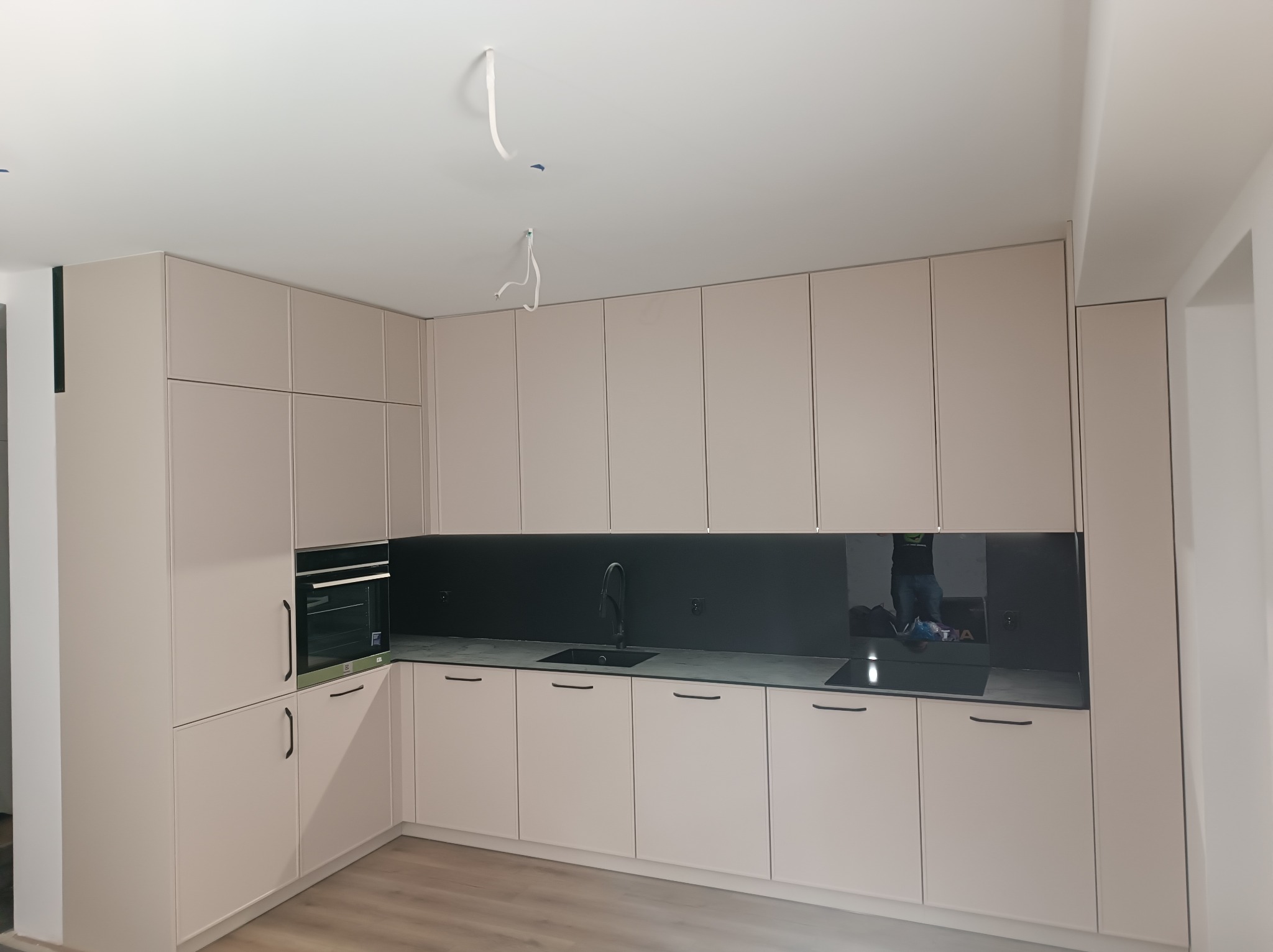 Jasna, nowoczesna kuchnia z beżowymi szafkami, czarnym blatem i panelem ściennym, widoczne niedokończone podłączenia elektryczne na suficie.