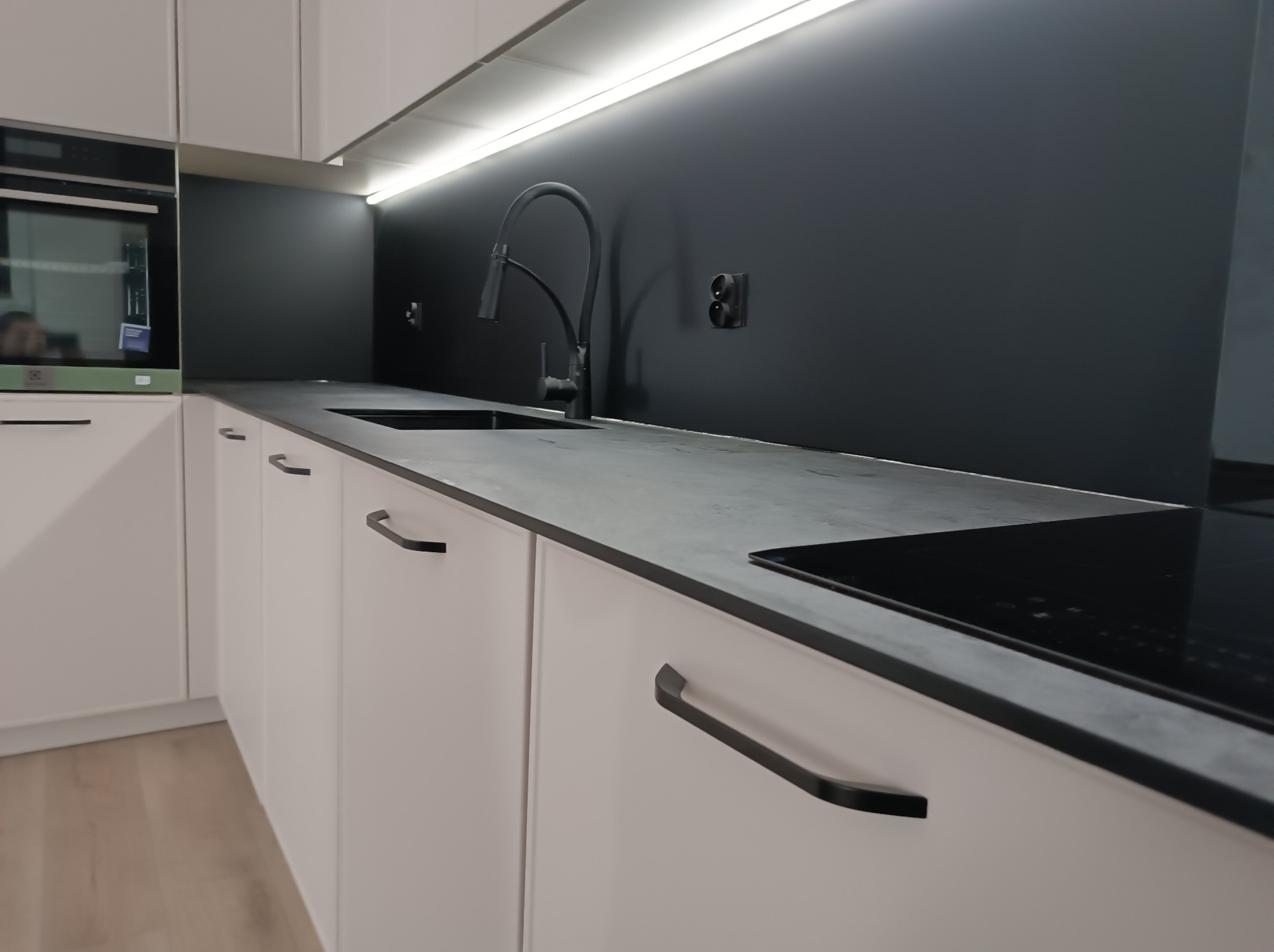 Nowoczesna, minimalistyczna kuchnia w Gorlicach: białe szafki z czarnymi uchwytami, szary blat, czarny panel nad blatem i oświetlenie LED pod szafkami wiszącymi.