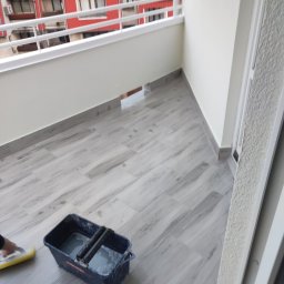 Kamil - Kładzenie płytek podłogowych na balkonie, widoczny fragment balkonu z wyłożoną podłogą z płytek imitujących drewno, wiadro z wodą i gąbką do czyszczenia fug, w tle budynki mieszkalne.