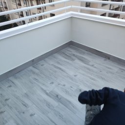 Kamil - Wykończony balkon w Łodzi z szarymi płytkami imitującymi drewno, białe ściany i balustrada, widok z góry z fragmentem osoby w ciemnej bluzie.