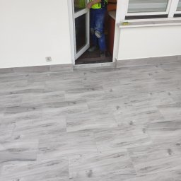 Kamil - Montaż drzwi wejściowych z widocznymi płytkami podłogowymi imitującymi drewno i fragmentem pracownika w odzieży roboczej.