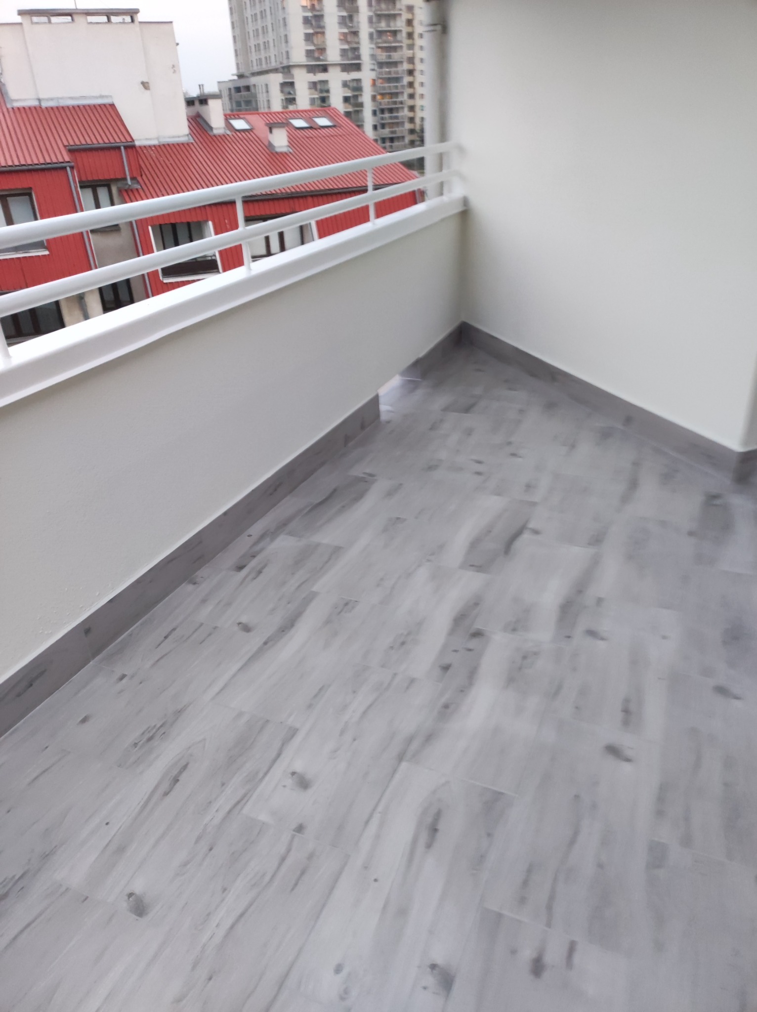 Wykończony balkon z szarymi płytkami imitującymi drewno, białe ściany i balustrada. Widok na budynki z czerwonymi dachami w tle.