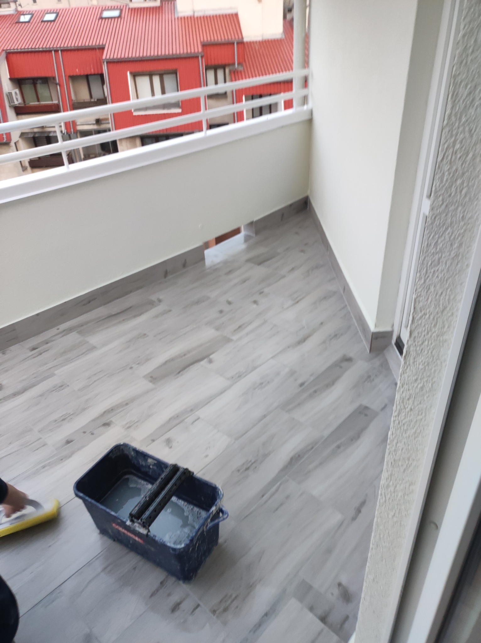 Kładzenie płytek podłogowych na balkonie, widoczny fragment balkonu z wyłożoną podłogą z płytek imitujących drewno, wiadro z wodą i gąbką do czyszczenia fug, w tle budynki mieszkalne.