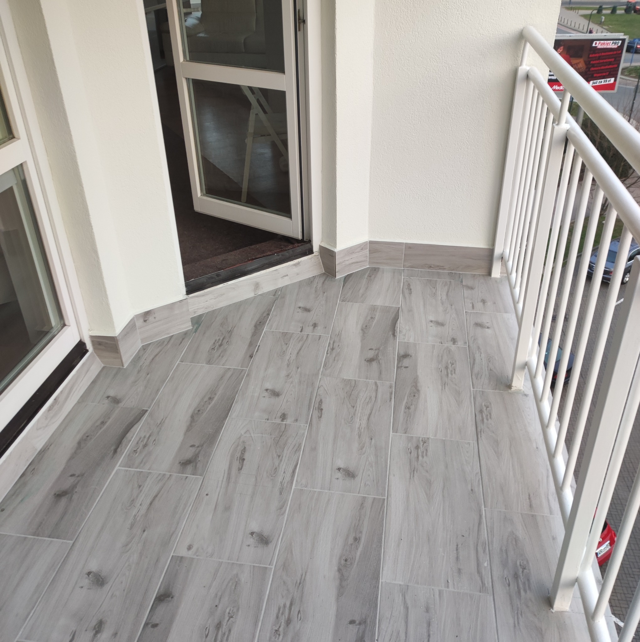 Wyłożony szarymi płytkami balkon z białymi barierkami i otwartymi drzwiami balkonowymi.