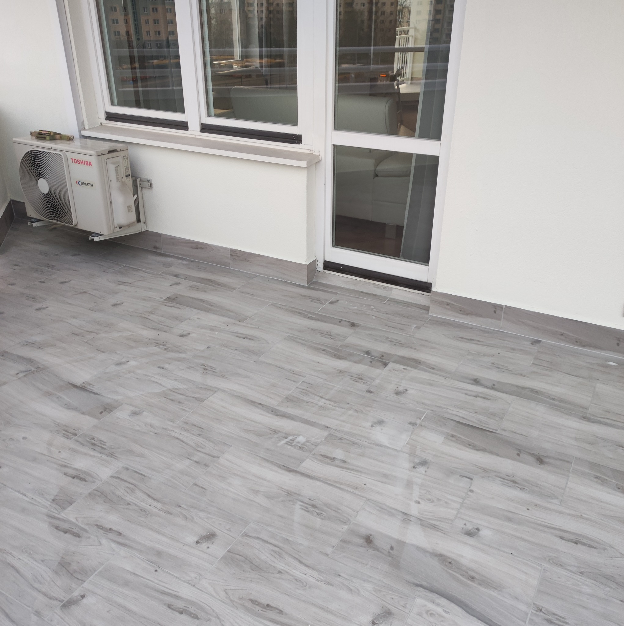 Wykończony balkon z szarymi płytkami imitującymi drewno, białe ściany i okno balkonowe, widoczny klimatyzator Toshiba.