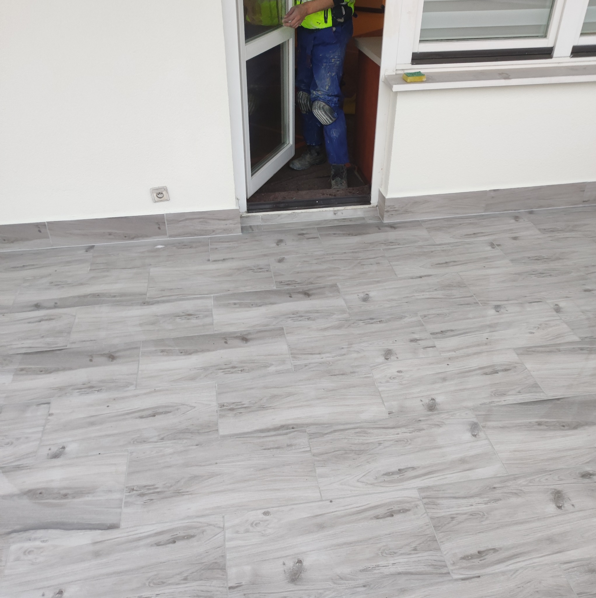 Montaż drzwi wejściowych z widocznymi płytkami podłogowymi imitującymi drewno i fragmentem pracownika w odzieży roboczej.