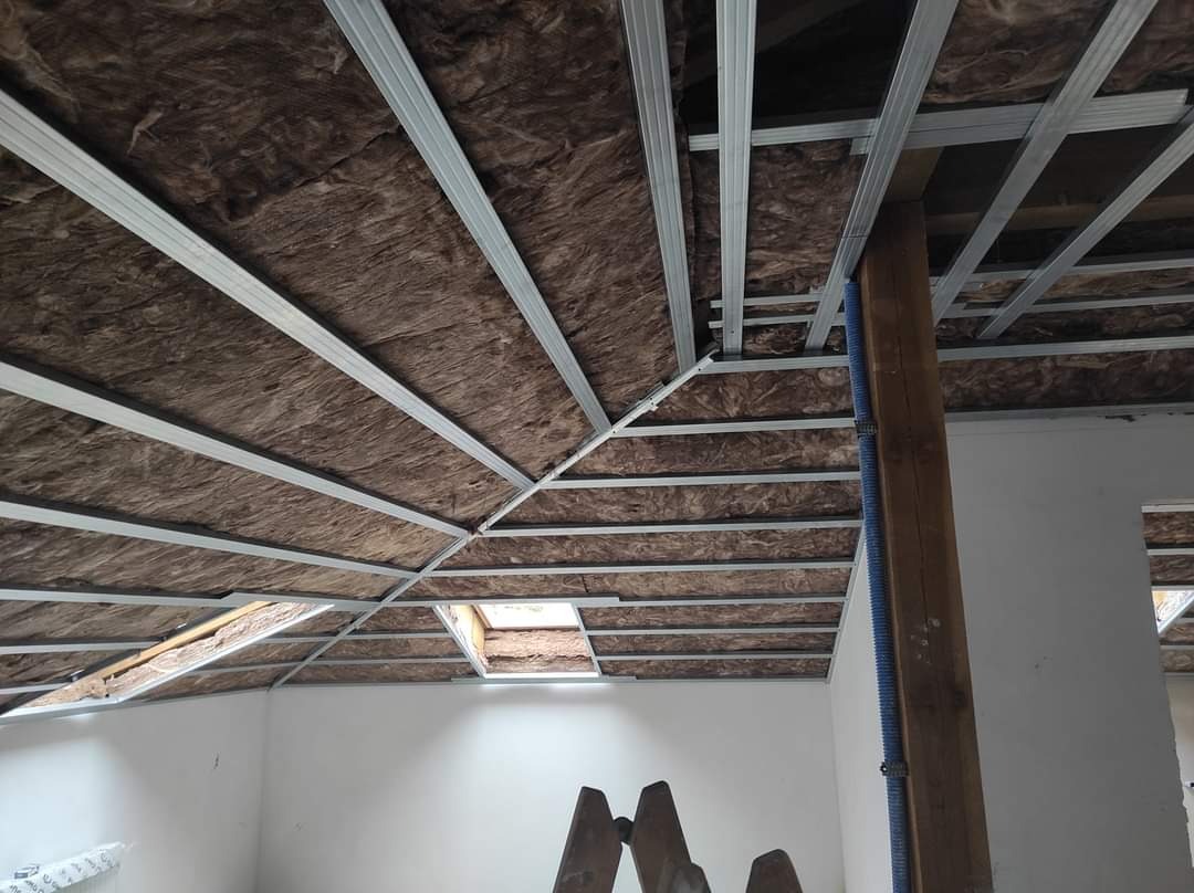 Poddasze z izolacją z wełny mineralnej i metalowym stelażem pod płyty gipsowo-kartonowe, widoczny fragment drewnianej kolumny z niebieskim przewodem oraz drabina.