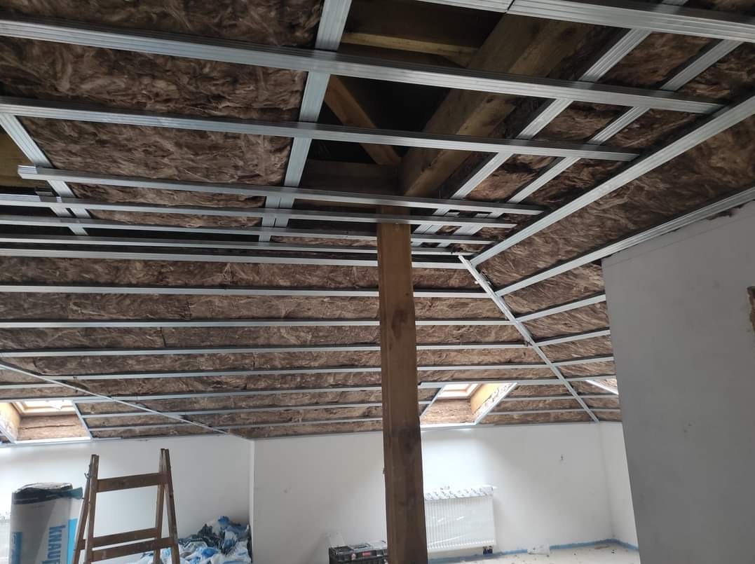 Sufit podwieszany z izolacją z wełny mineralnej, widoczna konstrukcja metalowa i drewniany słup podtrzymujący, w tle okno dachowe i grzejnik.