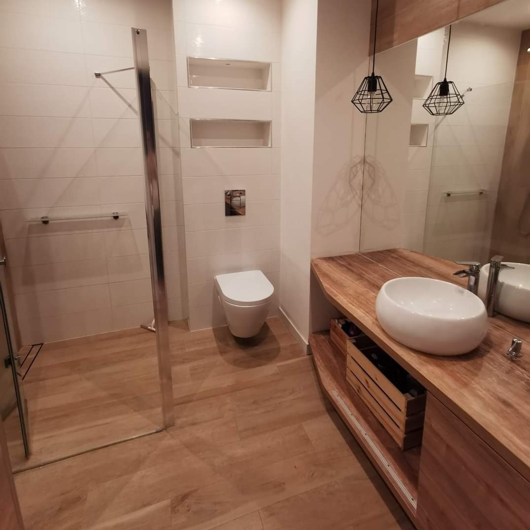 Nowoczesna łazienka z drewnianymi akcentami: kabina prysznicowa ze szklaną ścianą, umywalka nablatowa na drewnianym blacie, biała toaleta pod zabudowanymi półkami w ścianie.