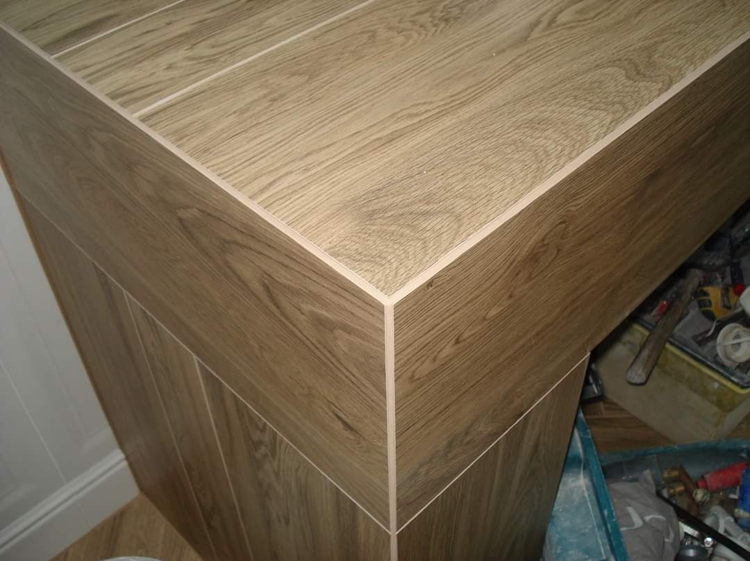 Wykończony narożnik obudowy w łazience z płytek imitujących drewno, widoczne narzędzia w tle.