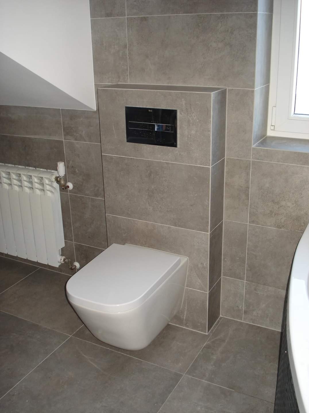 Nowoczesna łazienka z podwieszaną toaletą, szarą płytką imitującą kamień, białym grzejnikiem i czarnym panelem sterującym spłuczką.