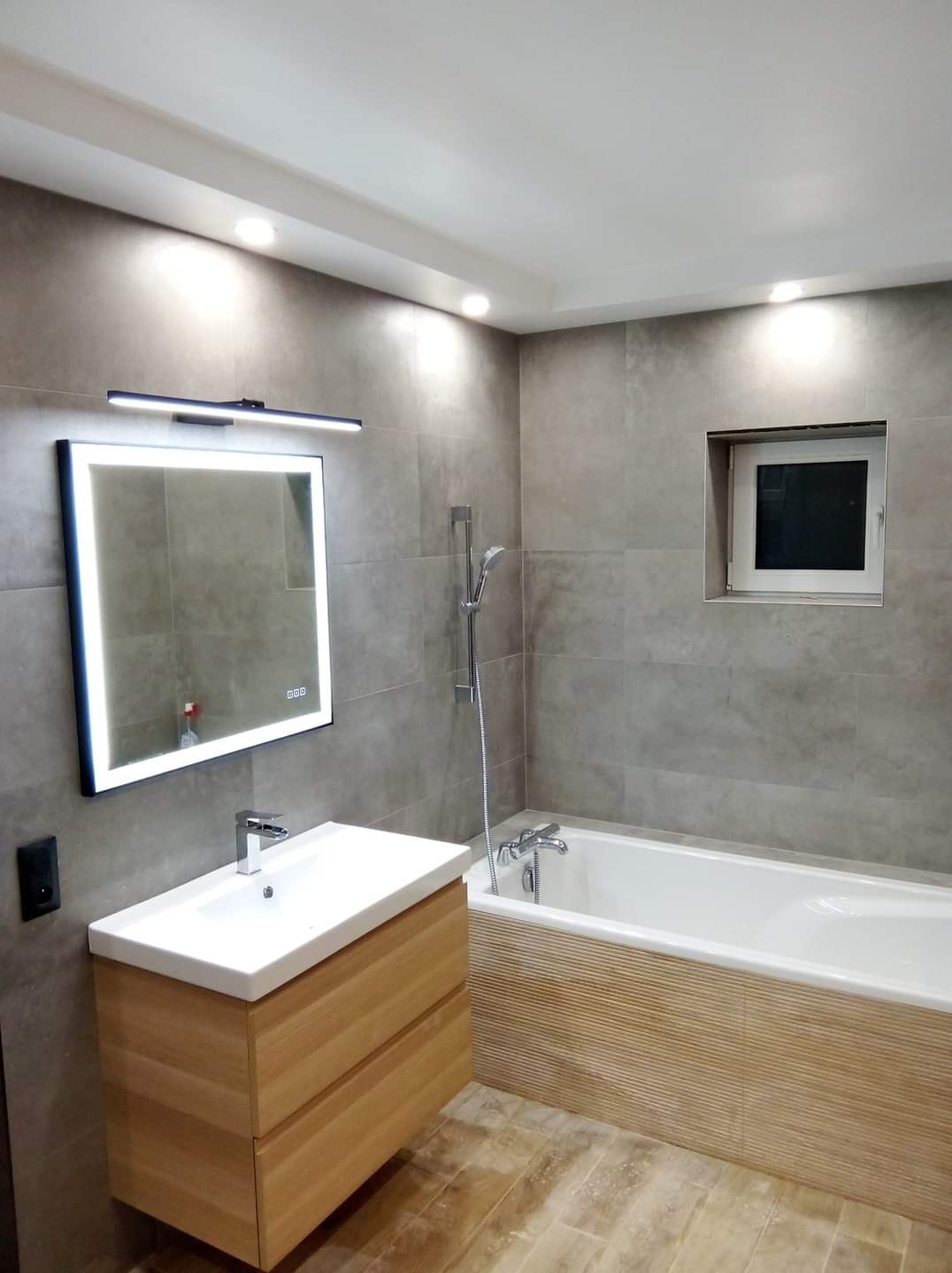 Nowoczesna łazienka z szarymi płytkami, drewnianą szafką pod umywalką, podświetlanym lustrem i wanną z dekoracyjnym panelem bocznym.