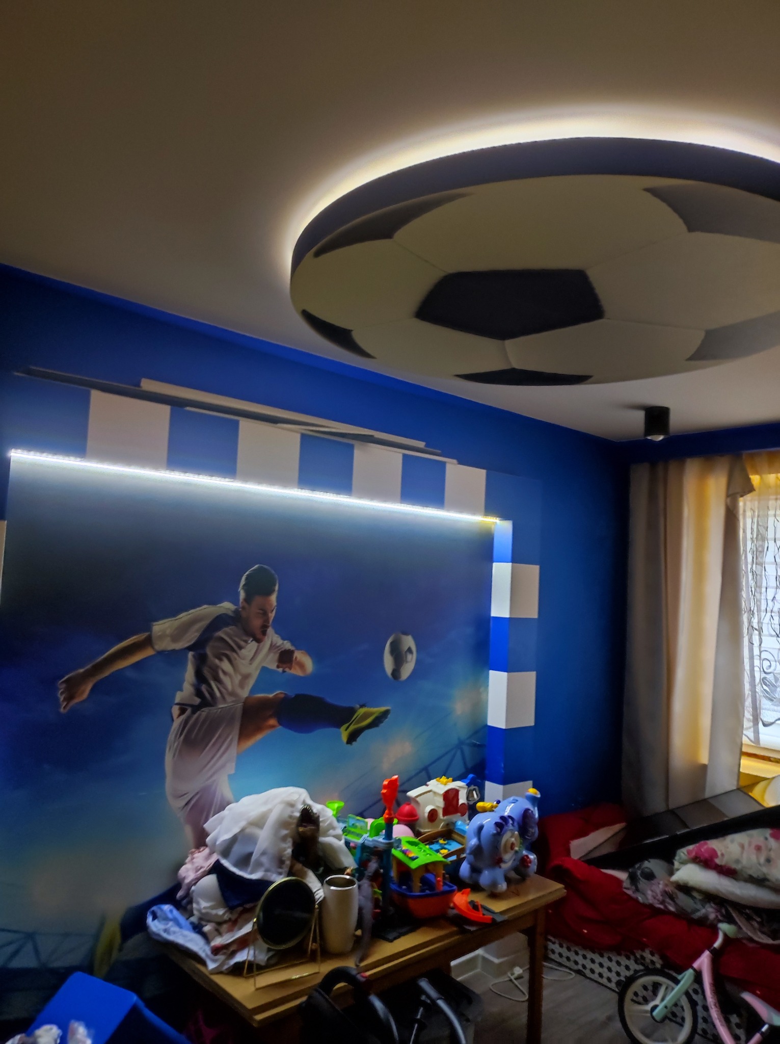 Pokój dziecięcy z fototapetą piłkarza, oświetleniem LED i lampą sufitową w kształcie piłki, widoczny lekki nieporządek na biurku i łóżku.