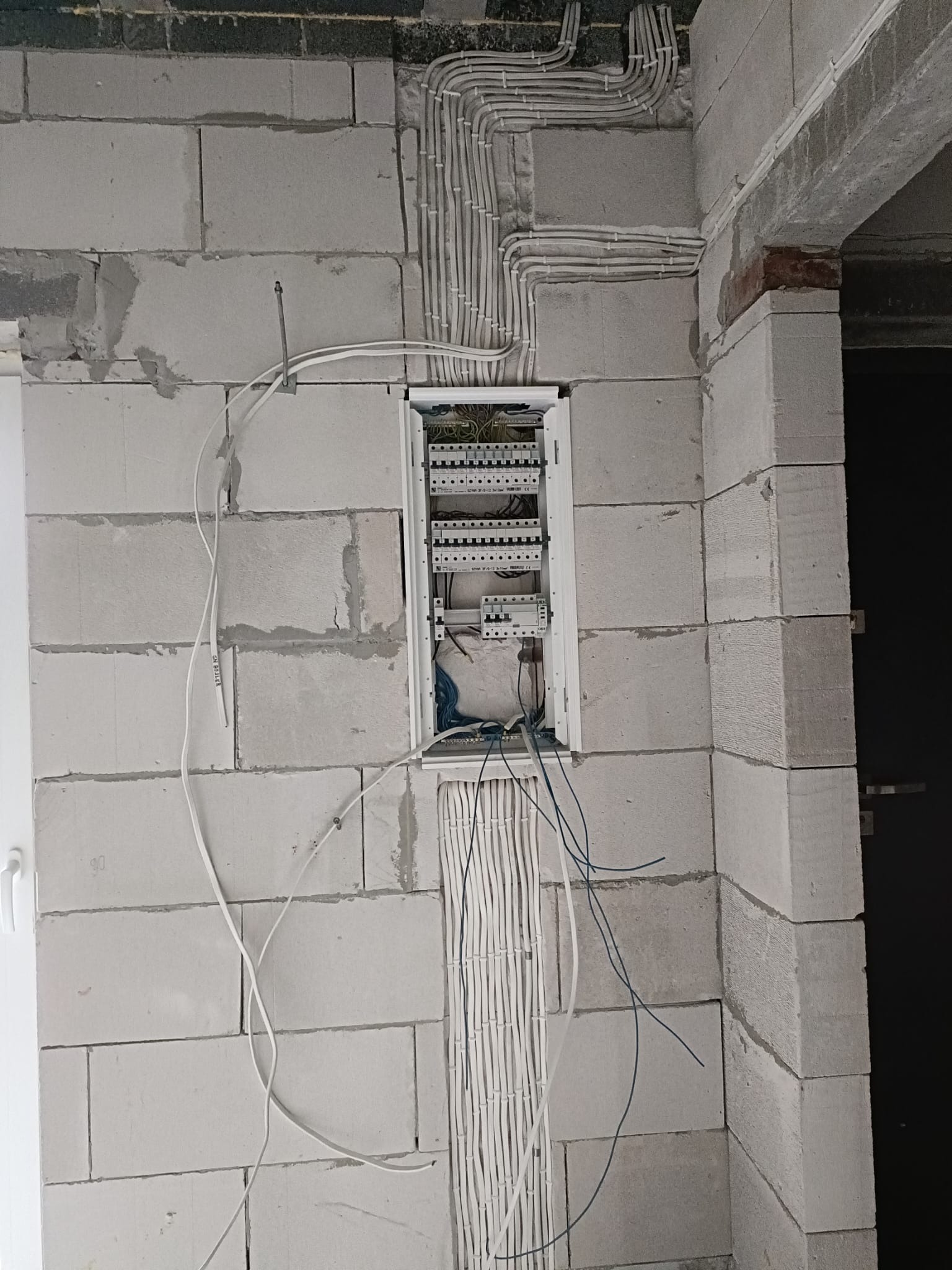 Rozdzielnia elektryczna w surowym stanie, kable prowadzone po ścianie z betonu komórkowego. Instalacja w trakcie montażu, widoczne zabezpieczenia i przewody.