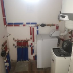 Wodavod - Rozdzielacz ogrzewania podłogowego z niebieskimi rurami, biały piec kondensacyjny i zmiękczacz wody w kotłowni z brązowymi płytkami podłogowymi.