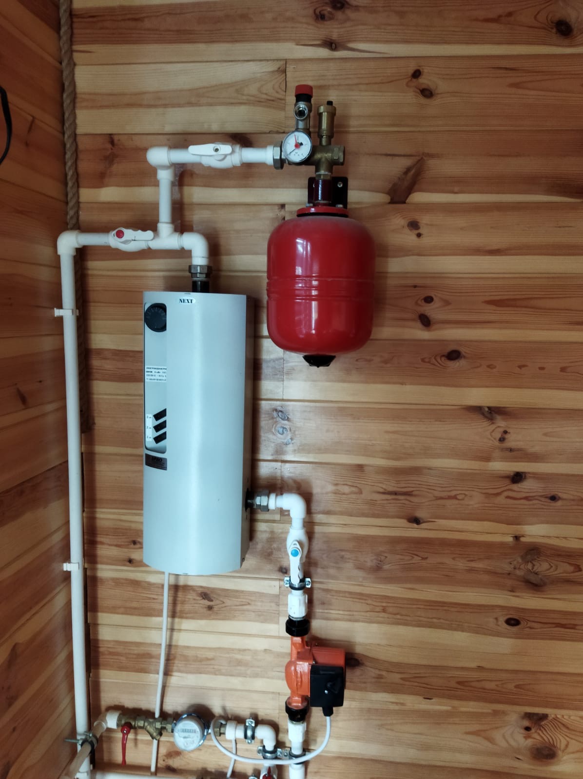 Montaż instalacji grzewczej z białym bojlerem, czerwoną przeponą i pompą obiegową na drewnianej ścianie.