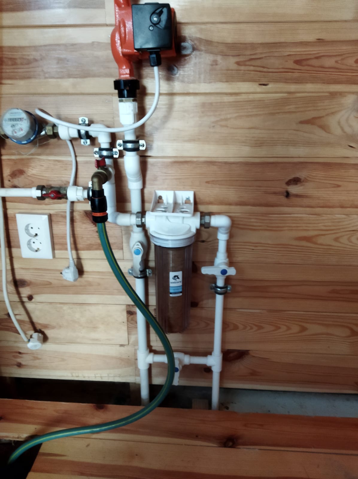 Instalacja wodna z filtrem mechanicznym, pompą obiegową i licznikiem, zamontowana na drewnianej ścianie w domku letniskowym.