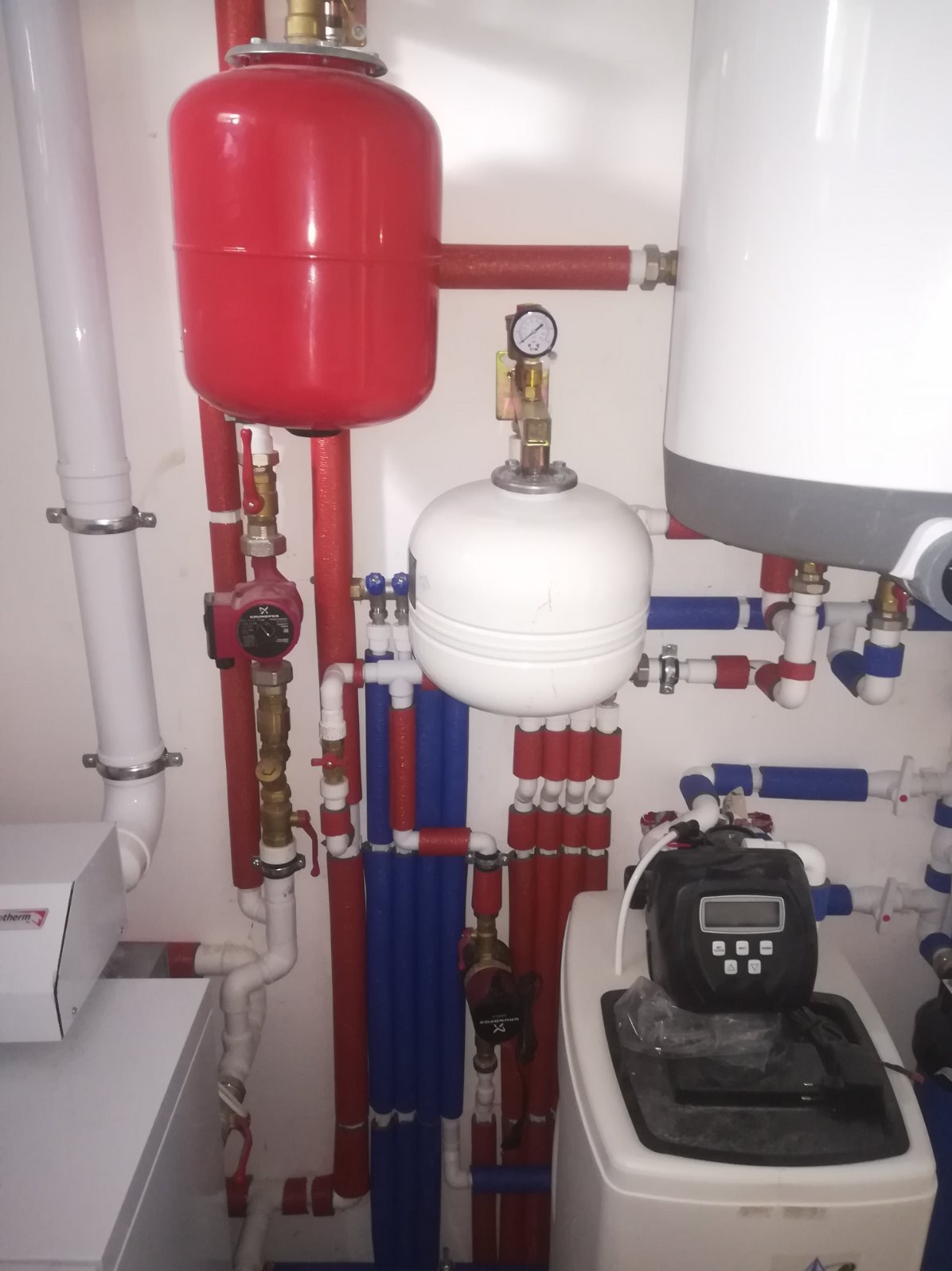 Instalacja kotłowni z czerwonym zbiornikiem wyrównawczym, białym zasobnikiem ciepłej wody, pompami obiegowymi i zmiękczaczem wody z elektronicznym wyświetlaczem, rury w izolacji czerwonej...