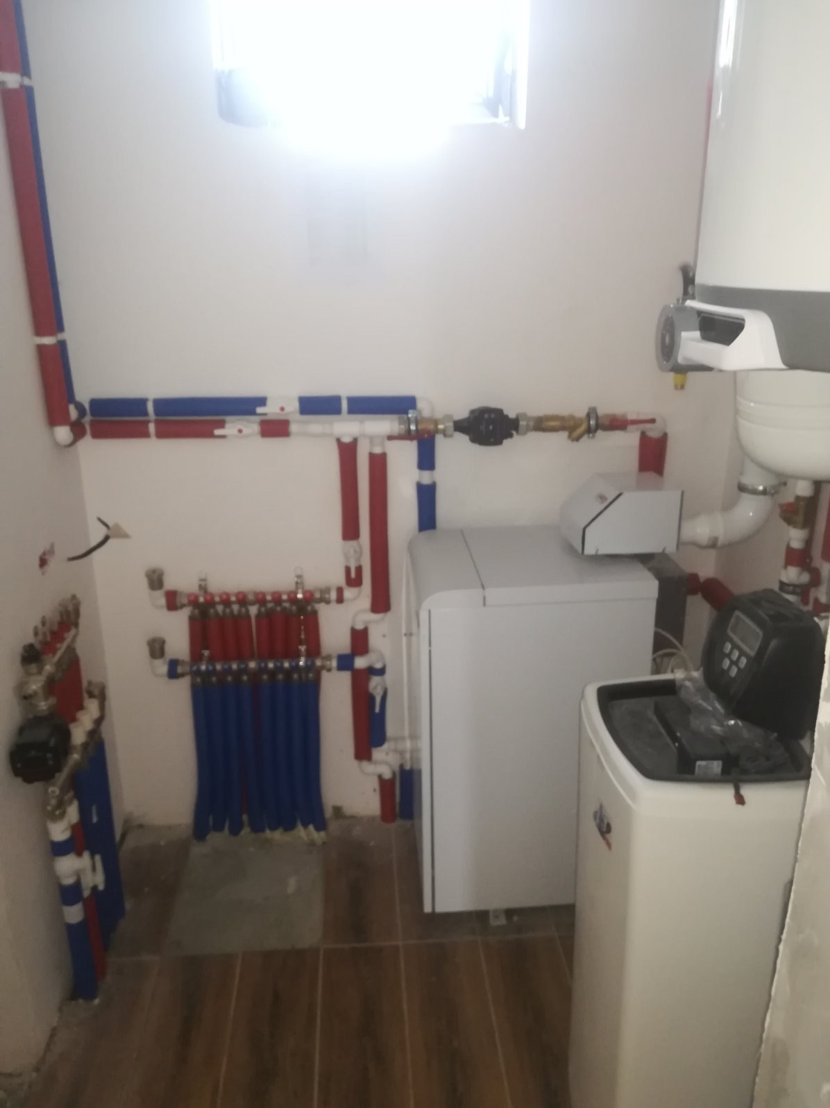 Rozdzielacz ogrzewania podłogowego z niebieskimi rurami, biały piec kondensacyjny i zmiękczacz wody w kotłowni z brązowymi płytkami podłogowymi.