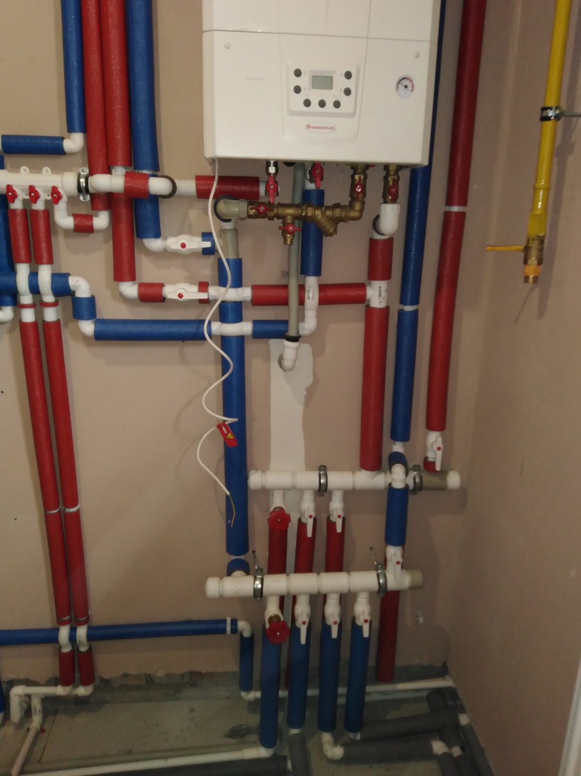 Biały piec gazowy Immergas z podłączeniami rur w izolacji czerwonej i niebieskiej, zawory z czerwonymi pokrętłami, żółty zawór gazowy, instalacja po montażu.