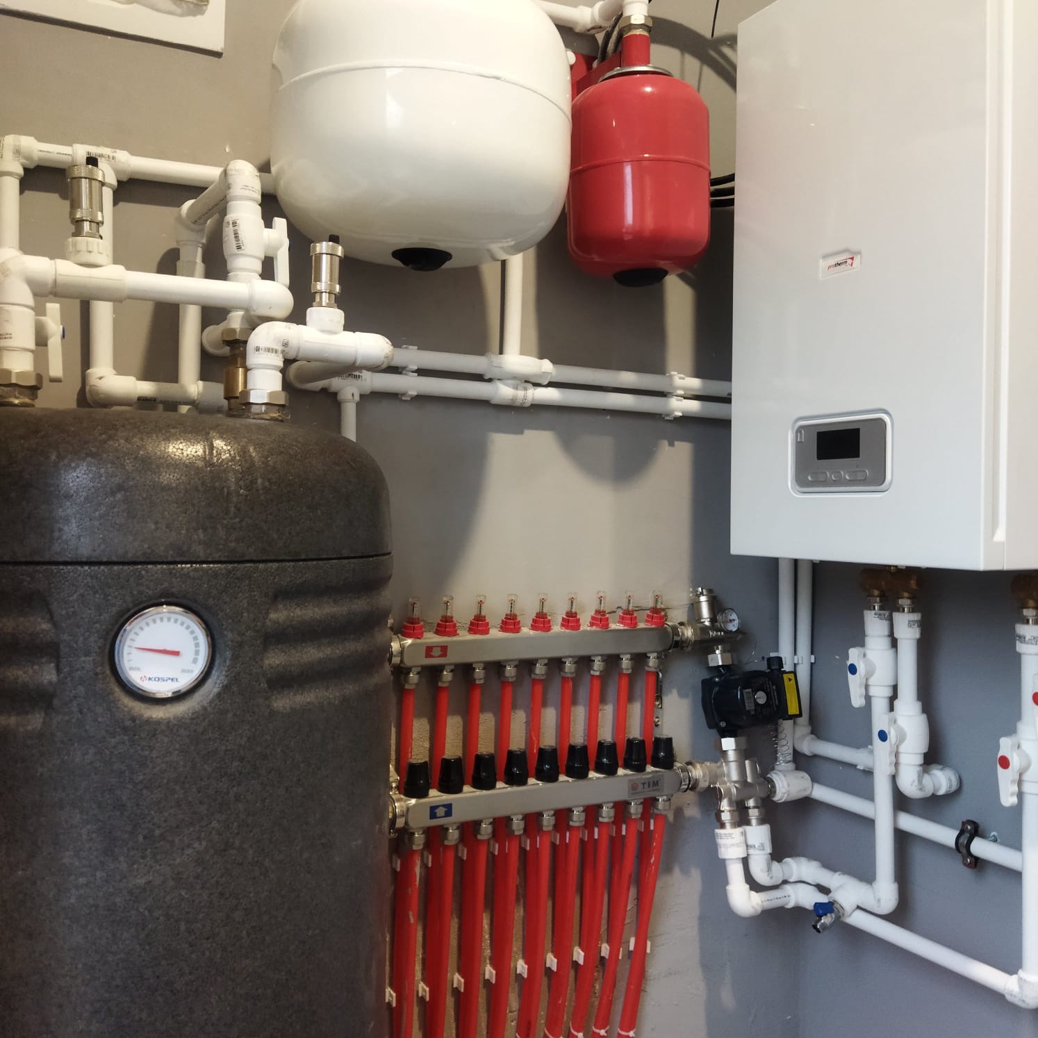 Zestawienie elementów instalacji grzewczej: piec, zasobnik, rozdzielacz ogrzewania podłogowego z licznymi rurkami, zbiorniki wyrównawcze, zawory i rury z tworzywa.