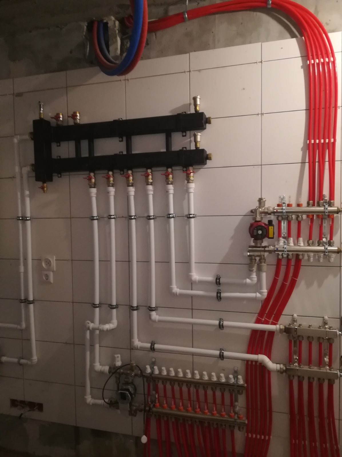 Rozdzielacze ogrzewania podłogowego z licznymi rurami, zaworami i mocowaniami na kafelkowanej ścianie w piwnicy.