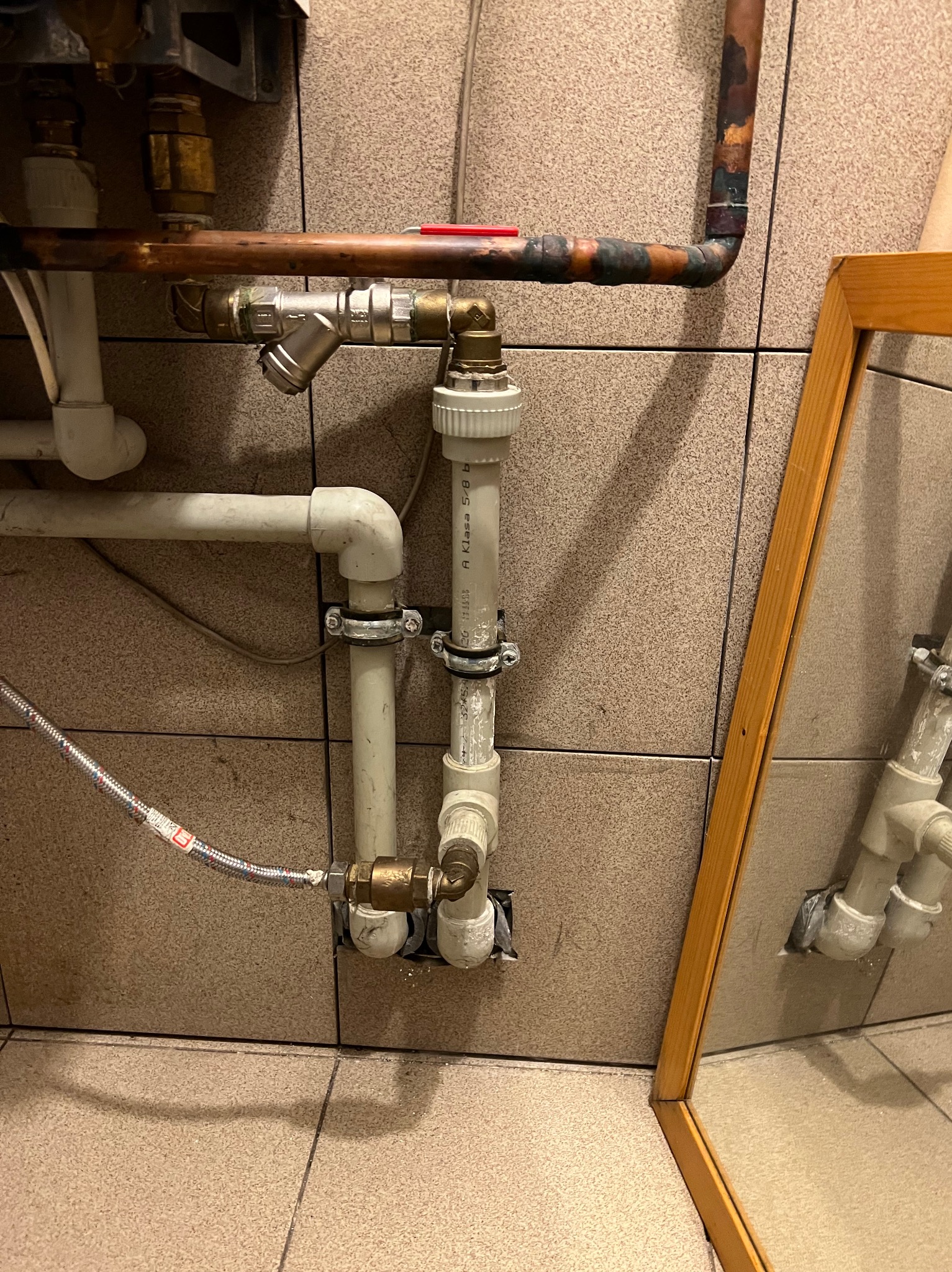 Połączenie rur miedzianych i plastikowych w łazience, widoczne zawory, złączki i fragment lustra w drewnianej ramie.