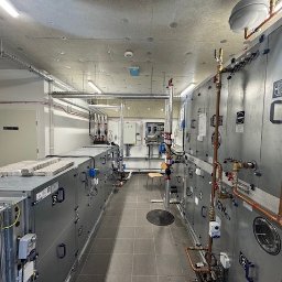 KLIMAZEN SPÓŁKA Z OGRANICZONĄ ODPOWIEDZIALNOŚCIĄ - Długi korytarz techniczny z dwoma rzędami szarych urządzeń wentylacyjnych i klimatyzacyjnych, widoczne rury miedziane, filtry na górze urządzeń po lewej stronie, krzesło w tle.