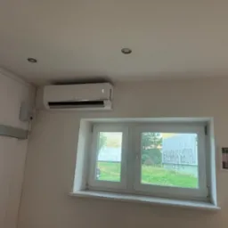 Biały klimatyzator LG zamontowany na ścianie nad oknem w pomieszczeniu, widoczne certyfikaty na ścianie obok okna.