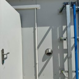 KLIMAZEN SPÓŁKA Z OGRANICZONĄ ODPOWIEDZIALNOŚCIĄ - Fragment ściany budynku z zamontowanym anemostatem, rurami instalacyjnymi i drabinką kablową, widoczne ostrzegawcze pasy na podłodze.