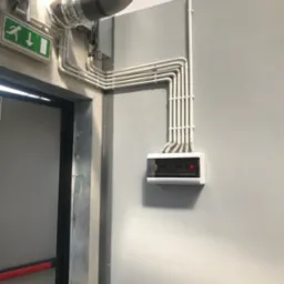 Szare pomieszczenie techniczne z instalacją wentylacyjną, rurami PCV i skrzynką elektryczną z wyświetlaczem LED, obok drzwi ewakuacyjnych z zielonym znakiem.