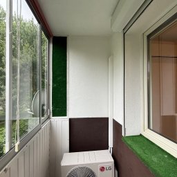 KLIMAZEN SPÓŁKA Z OGRANICZONĄ ODPOWIEDZIALNOŚCIĄ - Biała klimatyzacja LG Dual Inverter na balkonie z panelami PCV, sztuczną trawą i dużymi oknami. Widok na zieleń z zewnątrz.