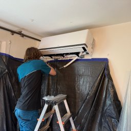 KLIMAZEN SPÓŁKA Z OGRANICZONĄ ODPOWIEDZIALNOŚCIĄ - Instalator montuje klimatyzator LG w pomieszczeniu zabezpieczonym folią ochronną. Widoczna drabina i przewody. Biały klimatyzator na jasnej ścianie.