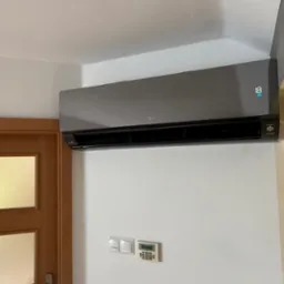 Szary klimatyzator LG, zamontowany na białej ścianie, obok drewnianych drzwi. Widoczny panel sterowania i wyświetlacz temperatury. Nowoczesny design.