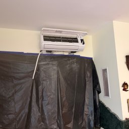 KLIMAZEN SPÓŁKA Z OGRANICZONĄ ODPOWIEDZIALNOŚCIĄ - Biały klimatyzator ścienny, otwarty, zamontowany nad czarną folią ochronną. Widoczne rury i kable. Ściana w kolorze kremowym, dekoracje na ścianie w tle.