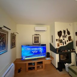 KLIMAZEN SPÓŁKA Z OGRANICZONĄ ODPOWIEDZIALNOŚCIĄ - Jasne wnętrze salonu z klimatyzatorem ściennym nad telewizorem. Na ścianach obrazy, kominek z dekoracjami. Widoczny fragment schodów.