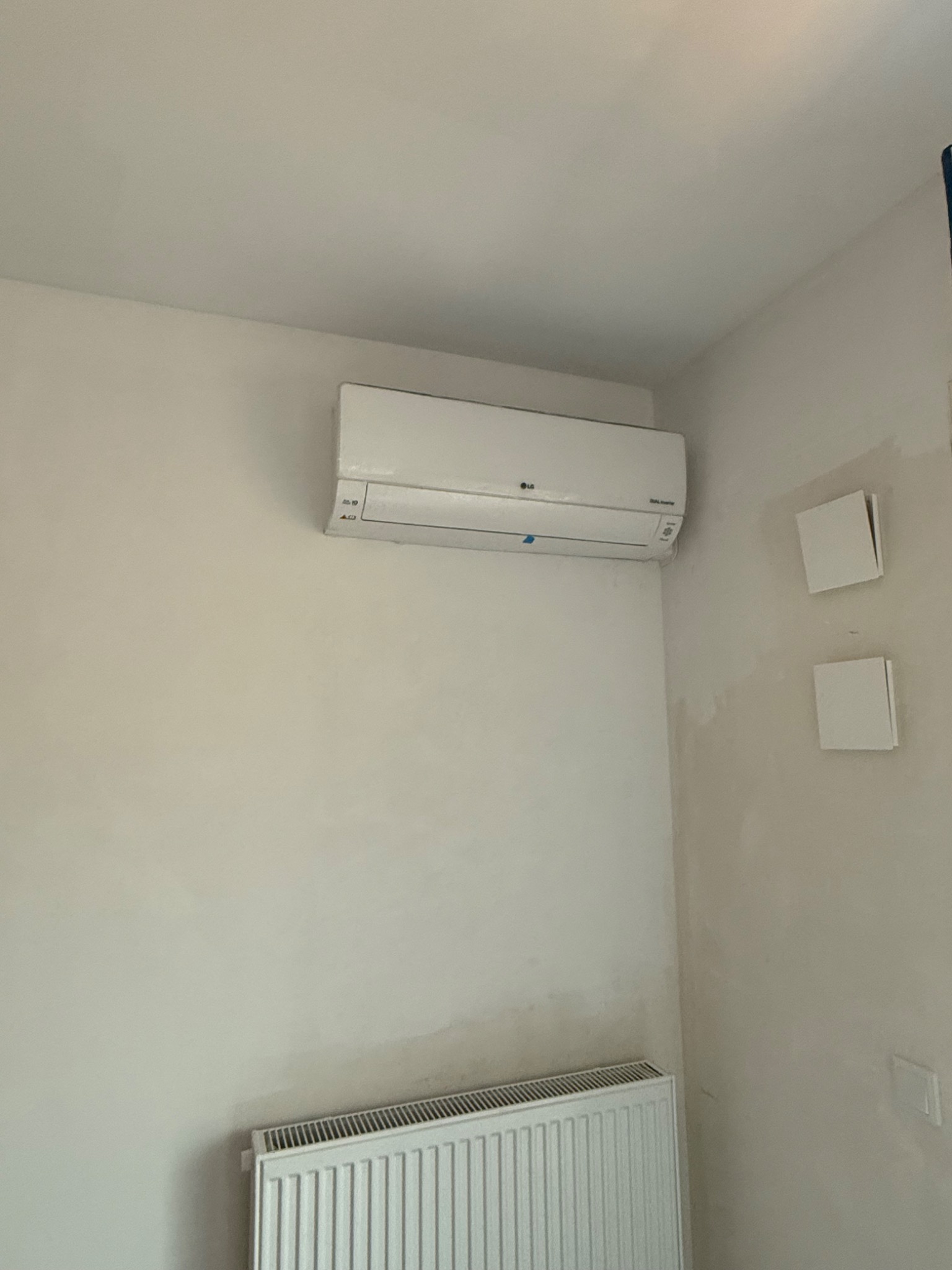 Biały klimatyzator LG zamontowany na ścianie w pokoju, widoczne kratki wentylacyjne i grzejnik.