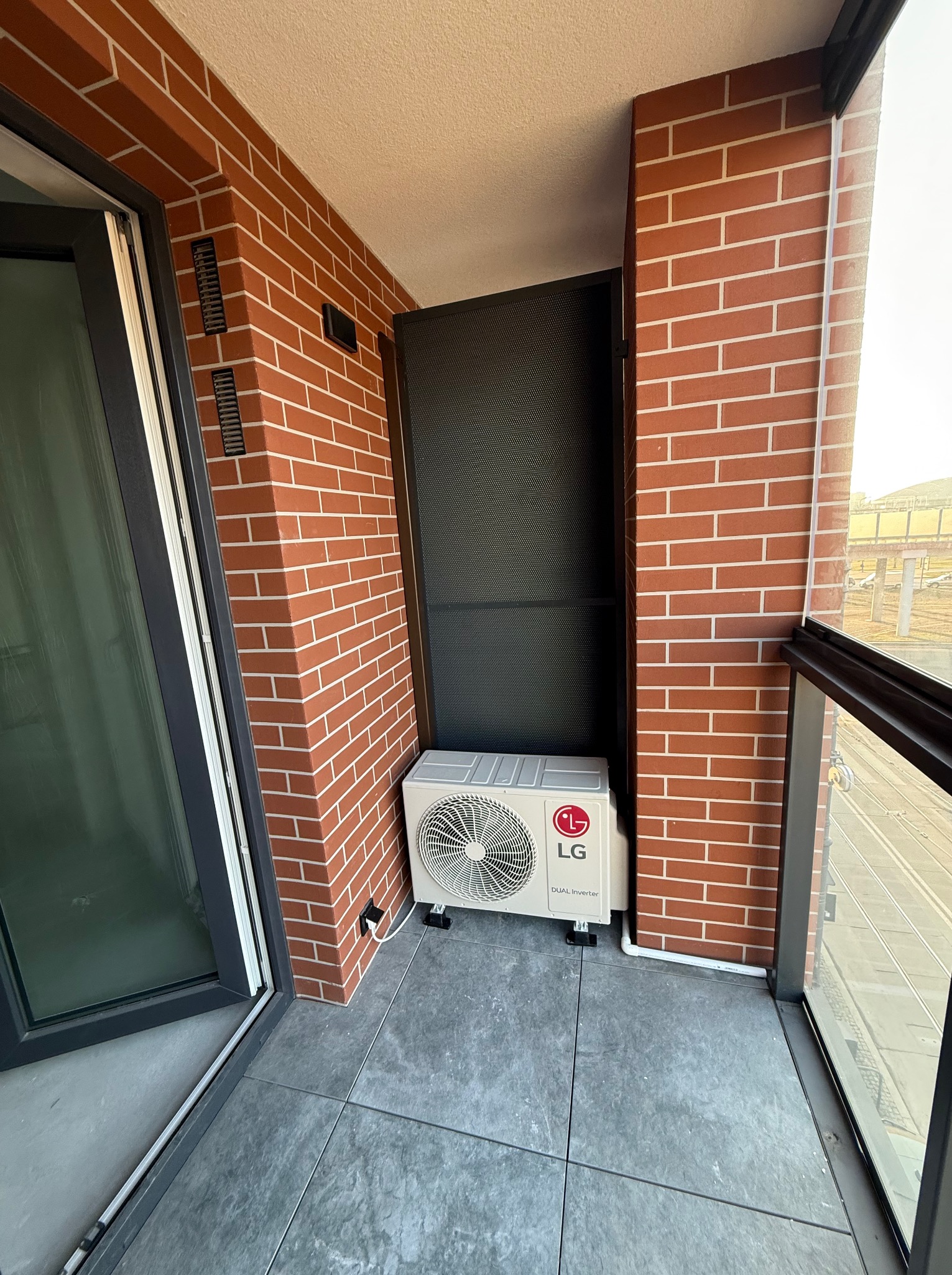 Kompaktowa jednostka zewnętrzna klimatyzatora LG Dual Inverter umieszczona na balkonie z ceglaną ścianą i szarą podłogą z płytek, widoczna rura odprowadzająca skropliny.