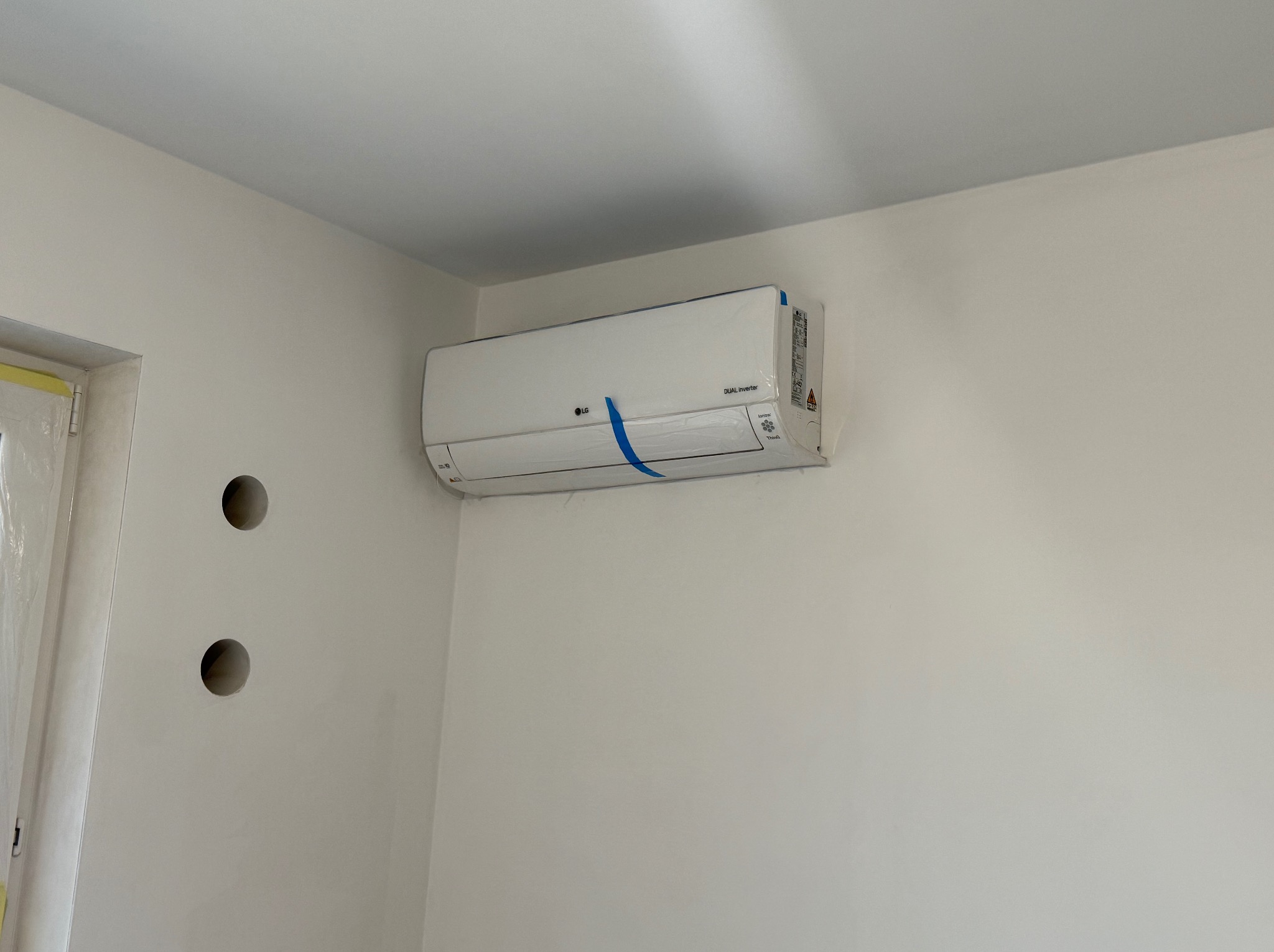 Nowy klimatyzator LG Dual Inverter, jeszcze w folii ochronnej, zamontowany na białej ścianie z widocznymi otworami na kable.