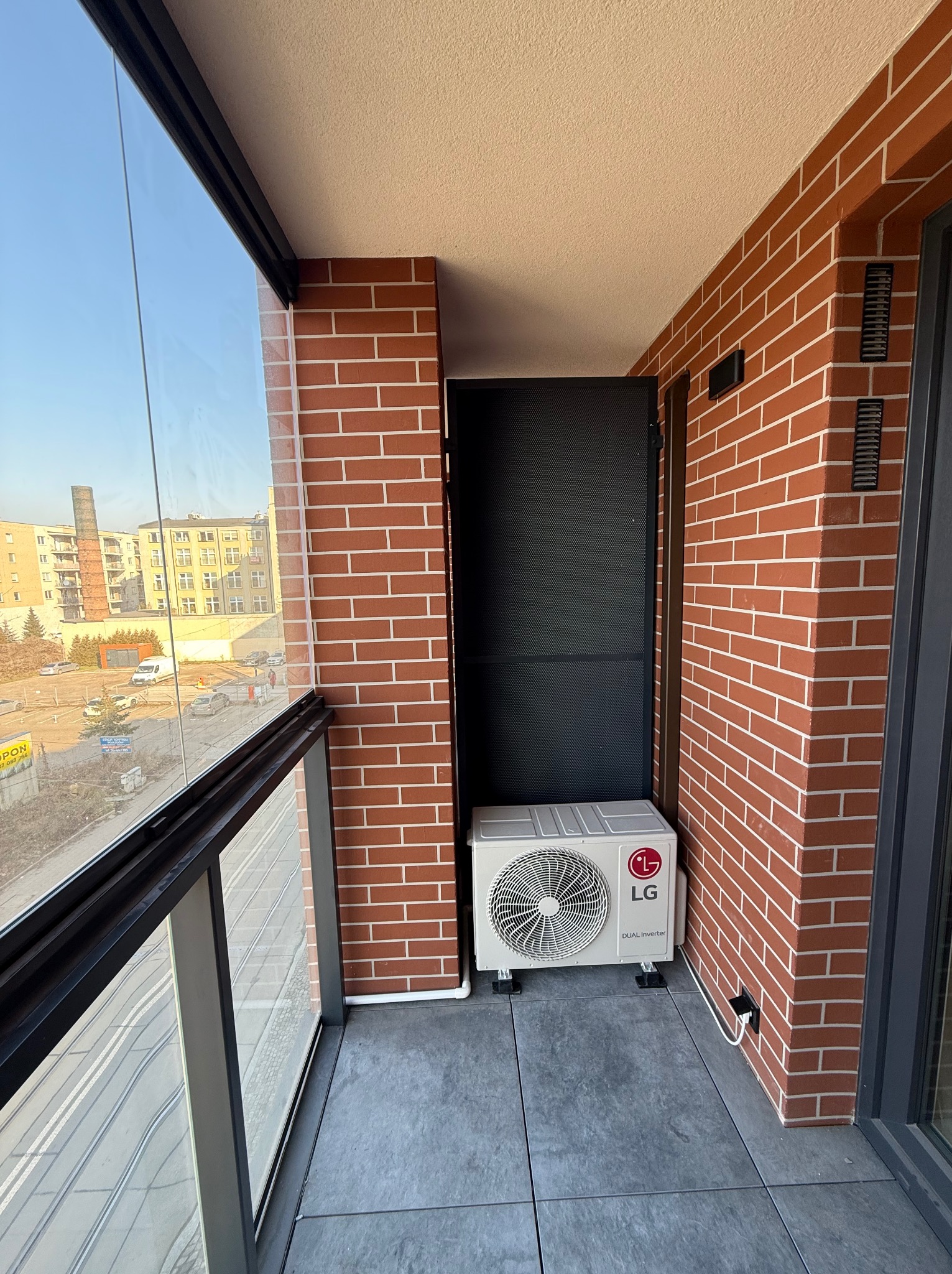 Biała jednostka zewnętrzna klimatyzacji LG Dual Inverter zamontowana na balkonie z ceglaną ścianą i widokiem na miasto za szklaną balustradą.